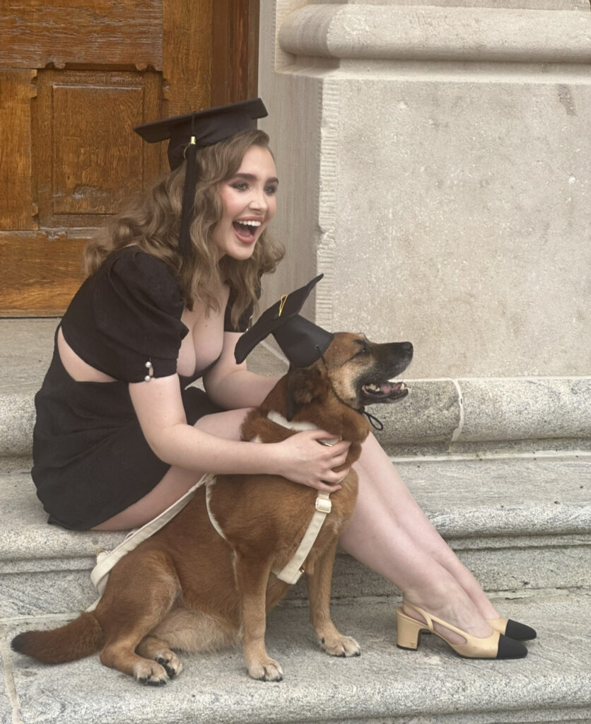 Kendyl sits on the steps of Seney Hall at Oxford College of Emory University in Oxford, Georgia. She sits with her dog, a small Chow Chow mixed breed, both wearing black and gold graduation caps.